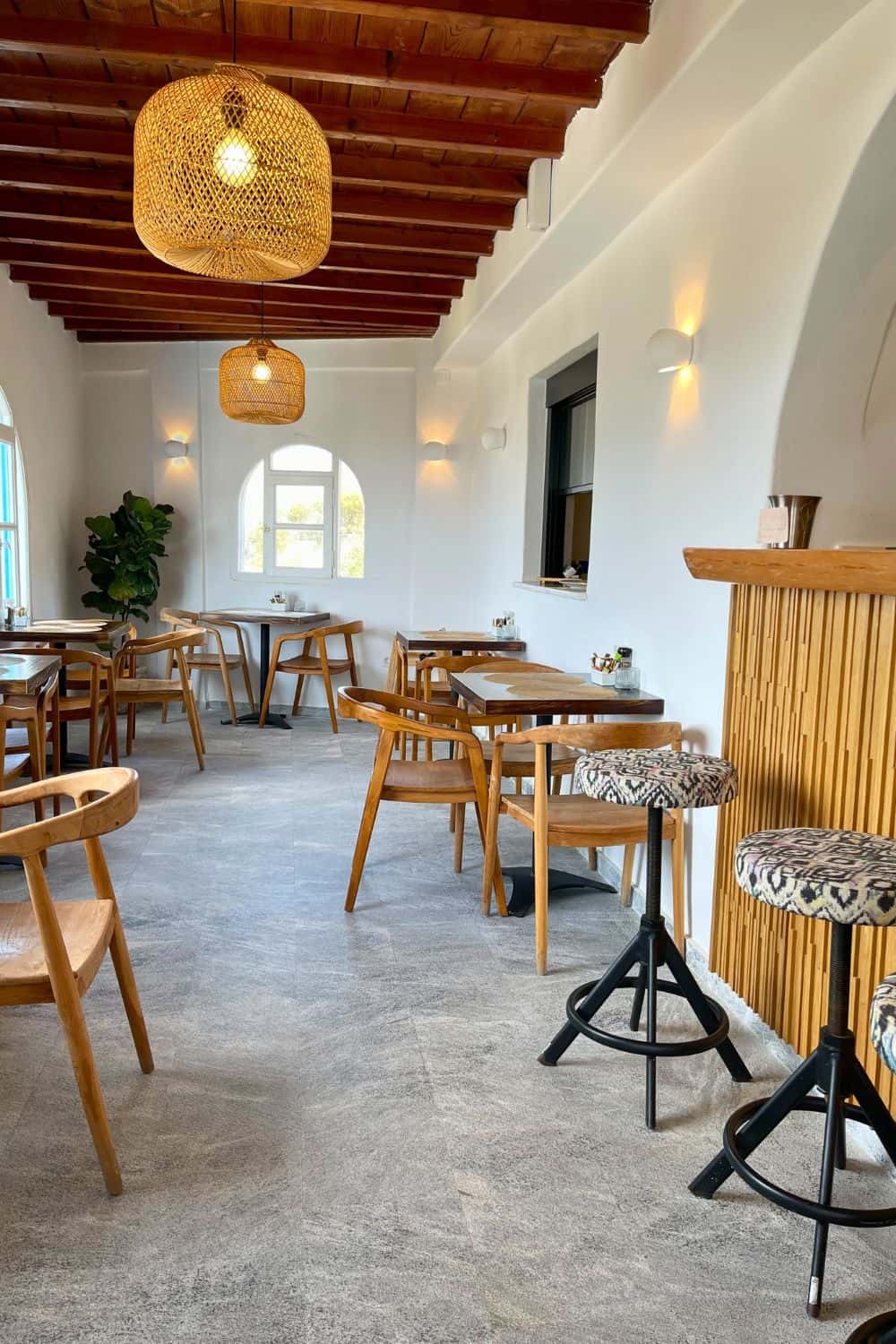 The interior of a cozy restaurant or café in Mykonos with a bright and airy feel. Wooden tables and chairs are neatly arranged on a grey tiled floor. The room is lit by natural light from windows and complemented by stylish hanging pendant lights with wicker coverings. The wooden ceiling beams add warmth to the space, creating a welcoming environment for diners