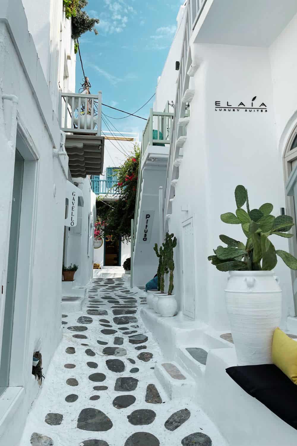 A picturesque narrow alley in Mykonos, framed by white Cycladic buildings with blue doors and windows. The path is adorned with smooth pebbles set in white plaster, creating a beautiful pattern that leads to a sign for 'ELAIA LUXURY SUITES,' indicating a boutique accommodation