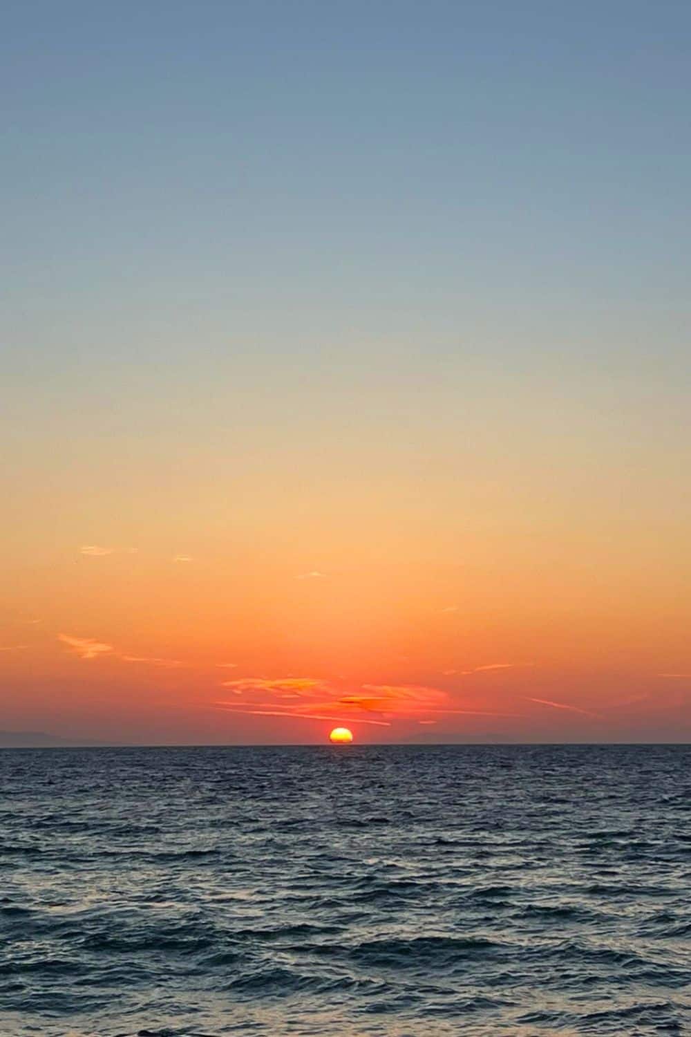 A serene sunset over the ocean with the sun dipping low on the horizon, casting a warm orange glow against the fading light of the sky. The gentle waves of the sea are capturing the last rays of the day, creating a tranquil and picturesque scene.