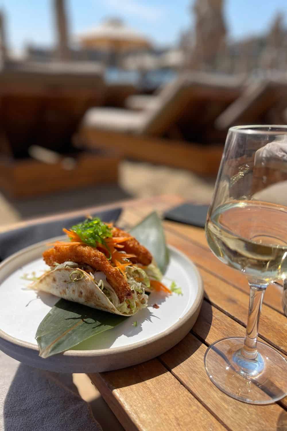 A close-up of a seafood taco on a white plate, paired with a glass of white wine on a wooden table with a blurred beach lounge background, capturing a relaxing dining experience by the sea.