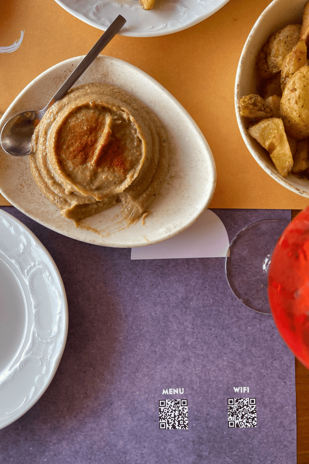 An overhead shot of a table at a restaurant featuring a plate of hummus with a dash of paprika on top, next to a bowl of roasted potatoes. A white plate, a glass with a red drink, and part of the restaurant's purple place mat with QR codes for a menu and WiFi are also visible