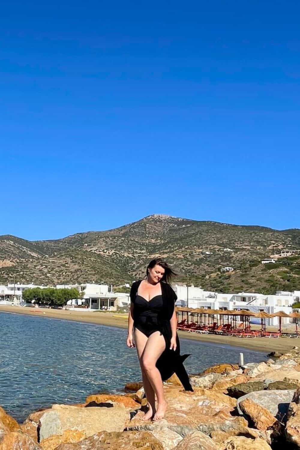 A woman in a chic black swimsuit poses gracefully on the rocky shore of a Sifnos beach, with the island's hilly landscape stretching out in the background under a clear sky, evoking the leisure and beauty of a seaside getaway.