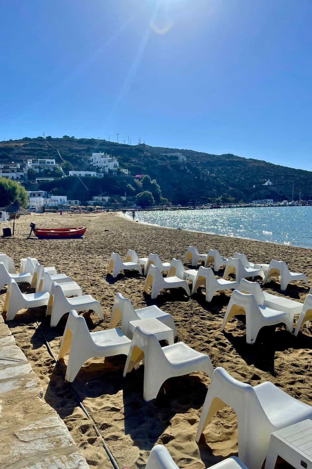 A serene morning scene on a Sifnos beach, with rows of white sun loungers waiting patiently for visitors. The calm blue sea complements the clear sky, while a traditional boat rests on the sand, highlighting the relaxed atmosphere of the island