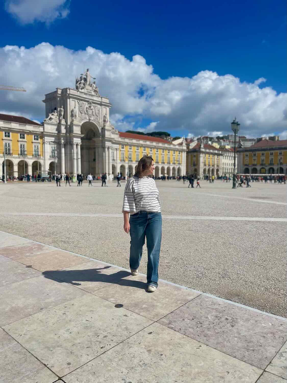Solo traveler walking towards the grand Rua Augusta Arch in the Praça do Comércio under a clear blue sky in Lisbon, Portugal.