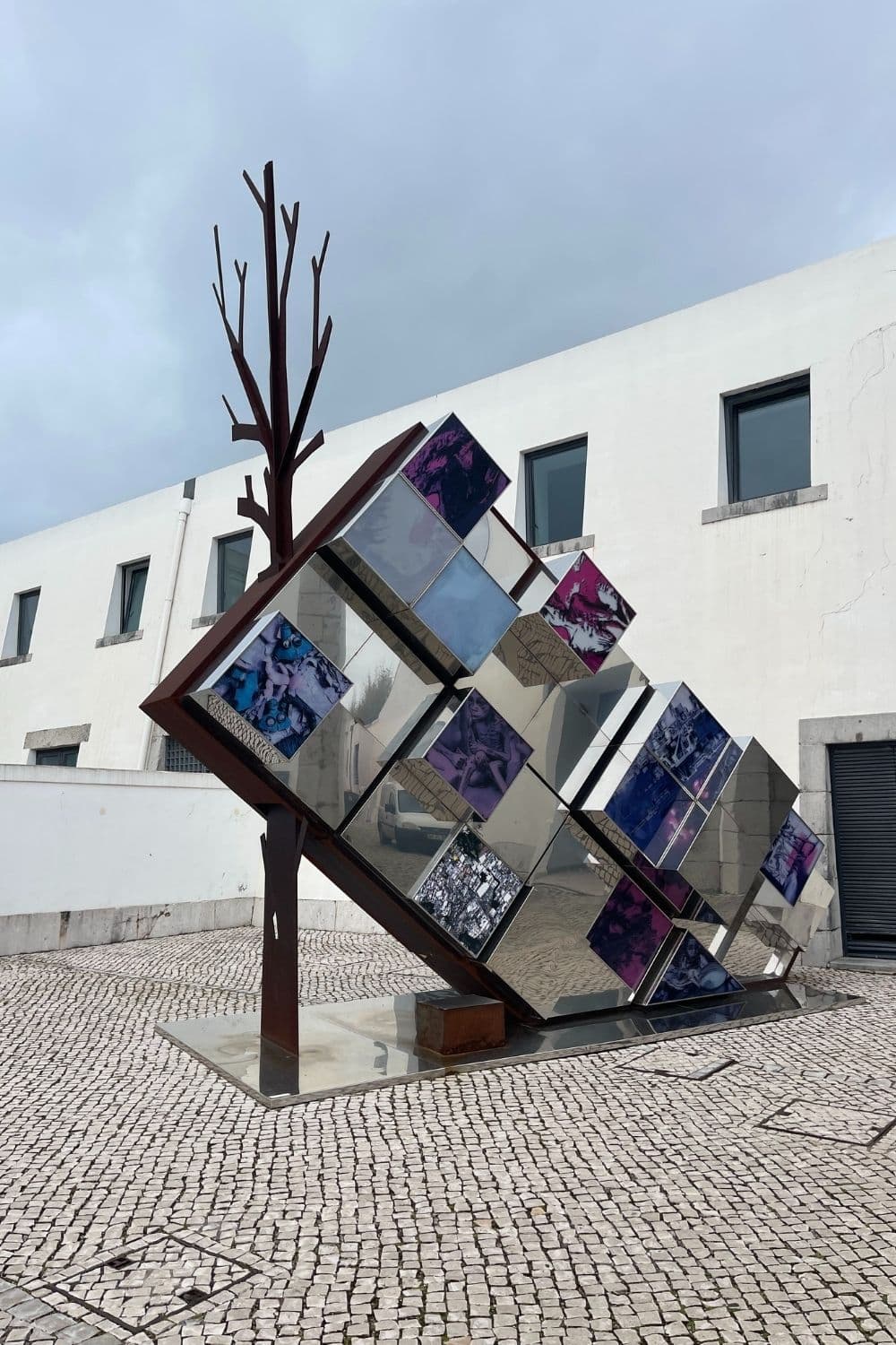An abstract outdoor sculpture featuring a tree-like metal structure with branches, adorned with mirrored, multi-angled panels reflecting various hues of blue and purple under an overcast sky, set against a backdrop of a white modern building with square windows.
