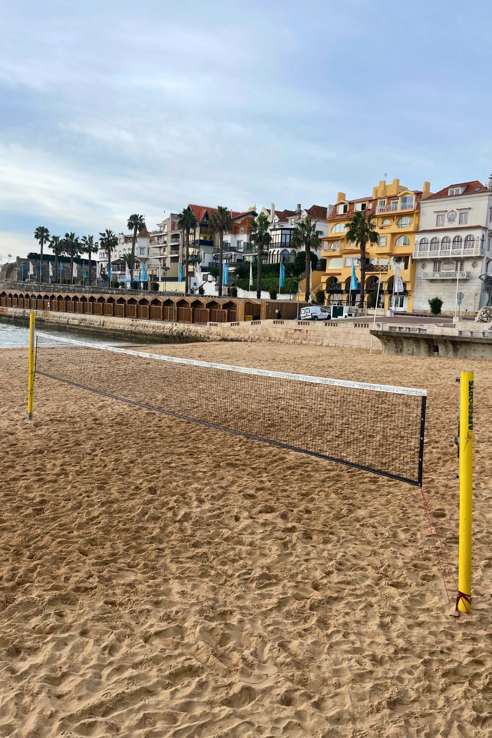 A sandy beach volleyball court awaits players in Cascais, with a net set against a picturesque backdrop of elegant houses and palm trees lining the promenade, under an overcast sky.