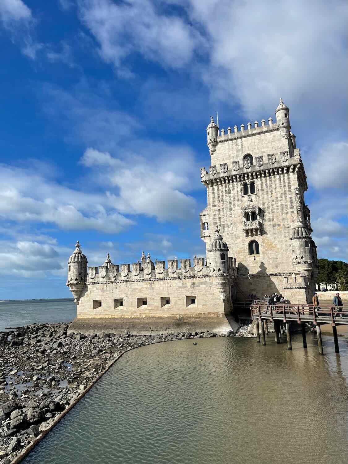 Belem Tower standing majestically beside the Tagus River under a partly cloudy sky, with a wooden footbridge leading to the entrance.
