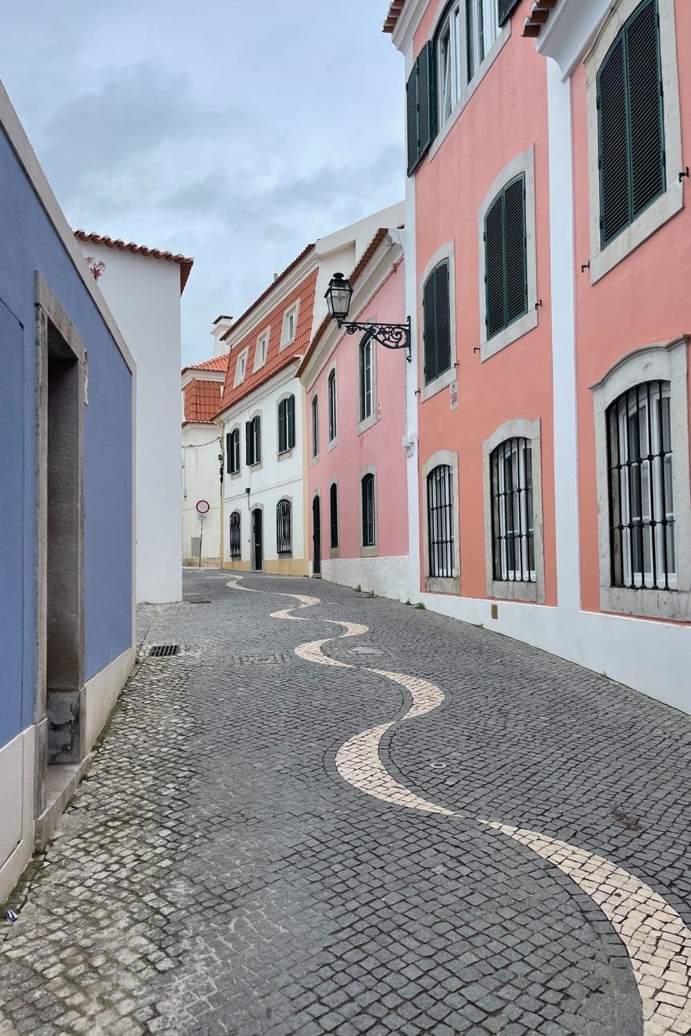 A quaint, curving cobblestone street lined with colorful pastel houses with traditional architecture, under a grey sky. The street features decorative, wave-patterned stone work and a vintage street lamp.