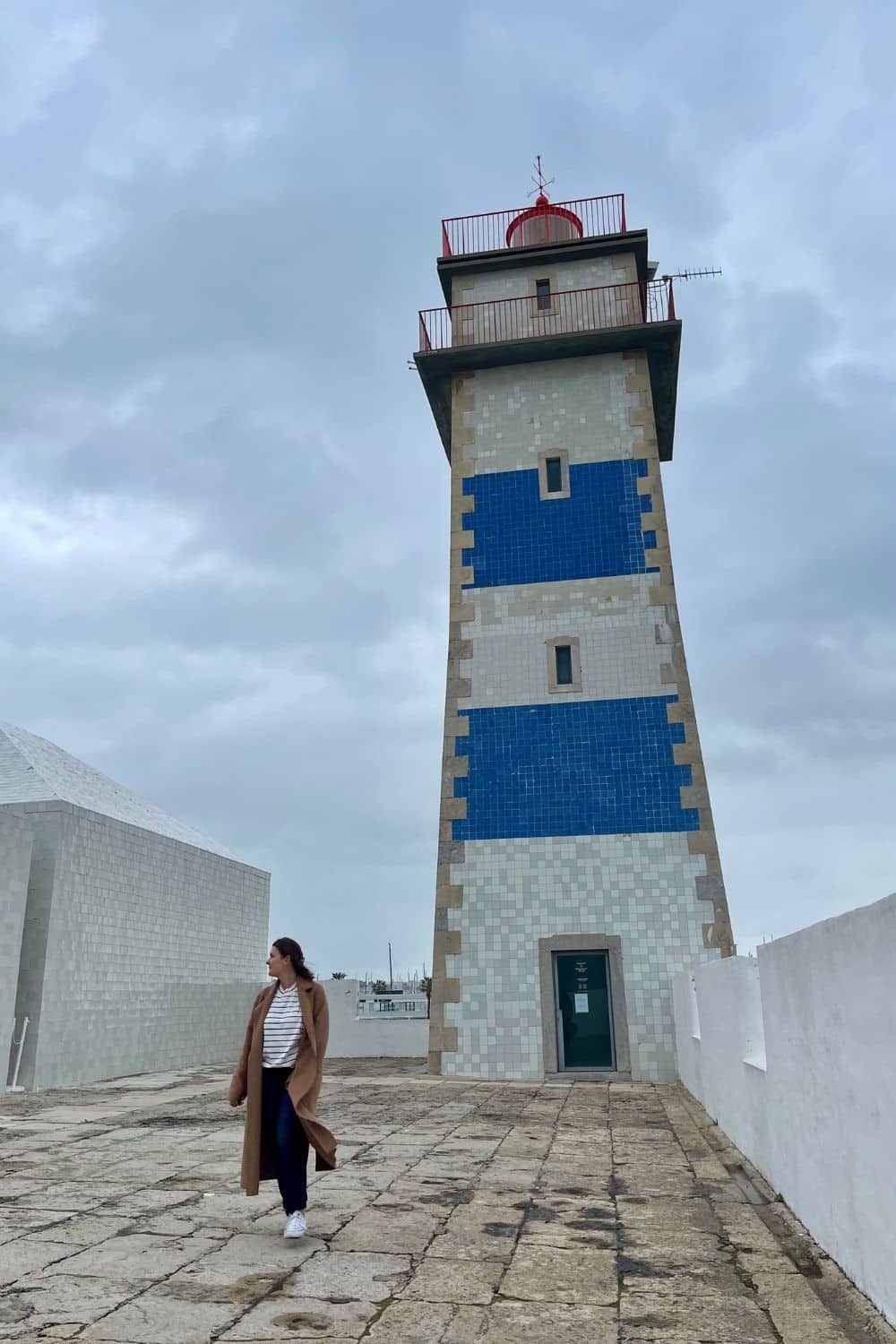 A woman in a long brown coat and white sneakers walks past a tall lighthouse with distinctive blue and white tiles, under a moody grey sky, highlighting the coastal heritage of Cascais