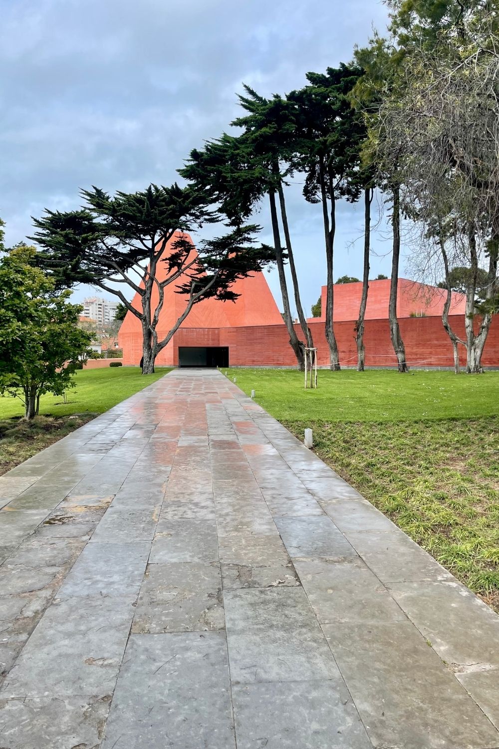 A pathway leading to the modern, terracotta-colored Paula Rego Museum in Cascais, Portugal, flanked by distinctive, windswept pine trees against a cloudy sky.