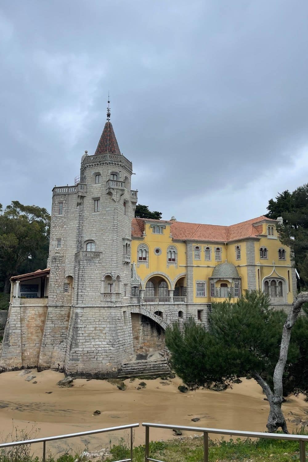 A whimsical yellow and white building with a stone tower and ornate features, reminiscent of a fairy-tale castle, set against a cloudy sky in Cascais, Portugal, conveying a sense of romantic history.