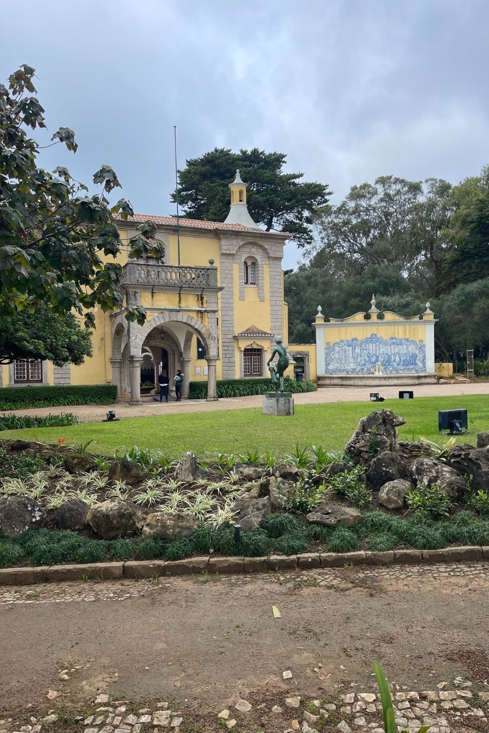 View of a historical yellow building with traditional Portuguese architecture, featuring azulejos (blue and white ceramic tiles) and a statue in the foreground garden, under a cloudy sky, capturing the essence of Cascais' cultural landscape.