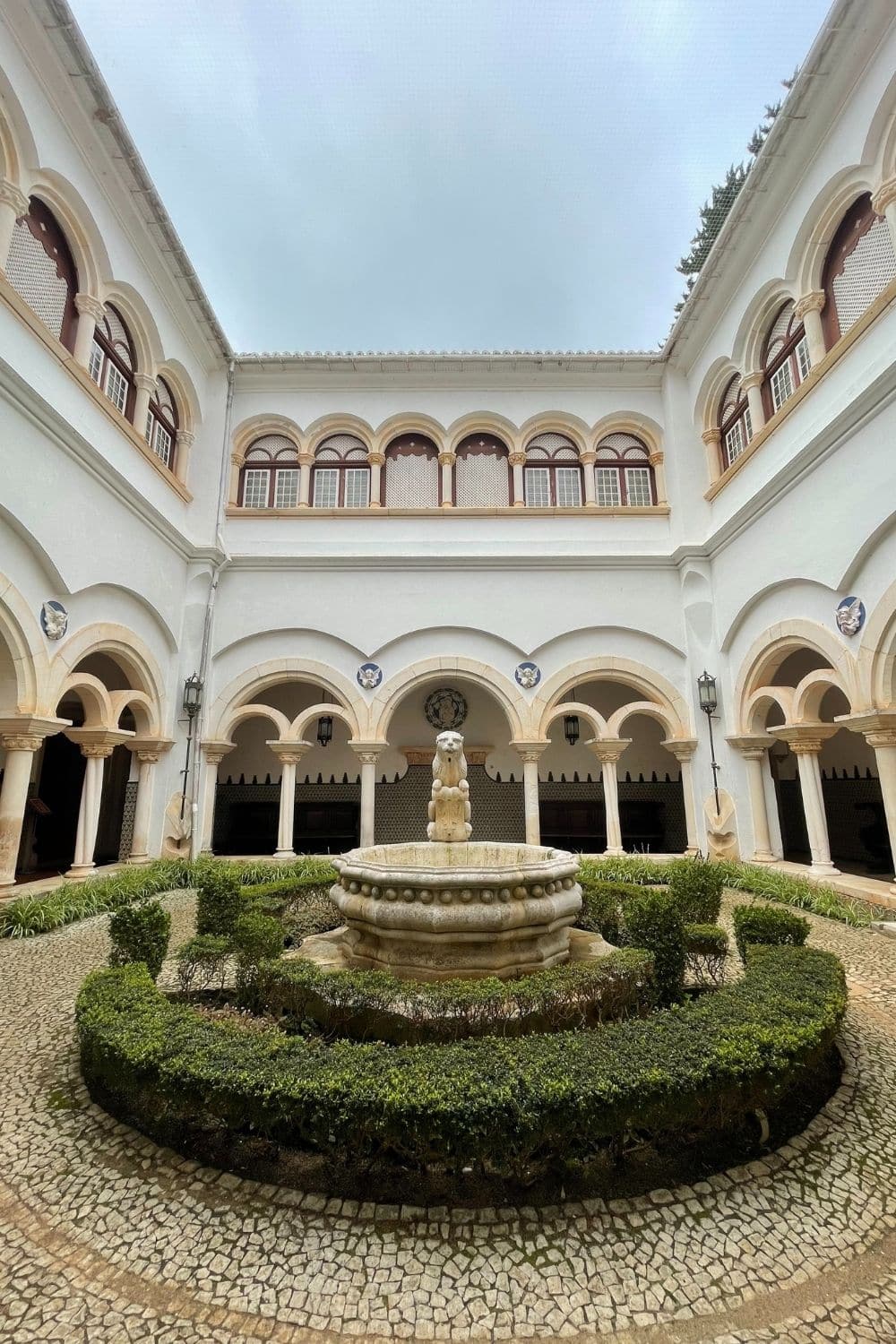 Elegant courtyard inside a historical building with a two-tiered fountain at its center, surrounded by archways and Moorish-style columns, under an overcast sky, reflecting Portuguese architecture.