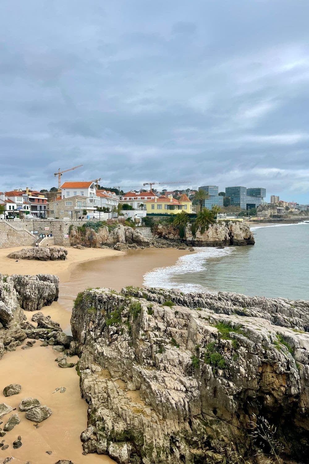 Scenic view of Cascais coastline with rugged rocks and a small sandy beach, contrasting with the urban backdrop featuring traditional and modern buildings under a cloudy sky, showcasing the town's natural and architectural beauty.