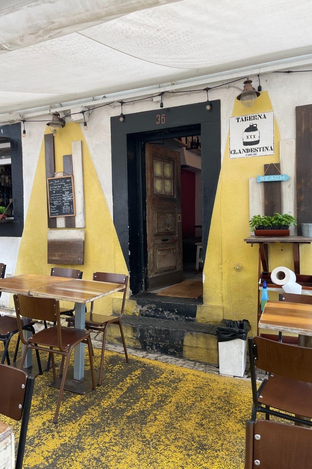 Exterior of Taberna Clandestina, with a yellow facade and black trim around the entrance, featuring rustic wood and metal furniture on cobblestone sprinkled with fallen yellow leaves, welcoming diners in Cascais.