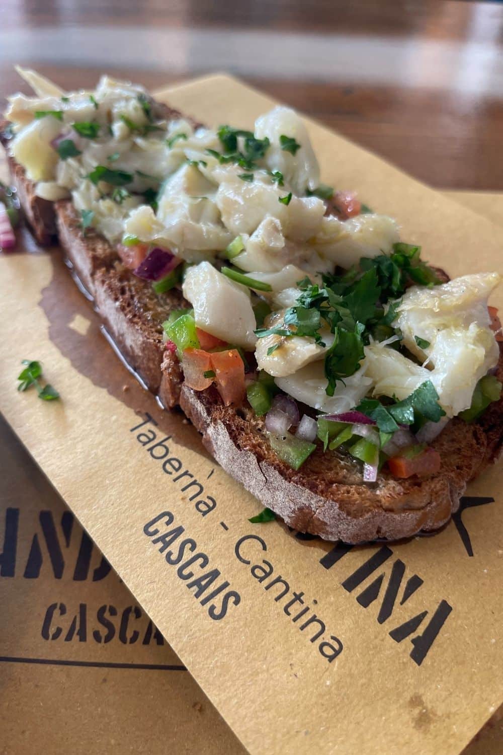 Artfully presented cod bruschetta on a rustic slice of bread, topped with diced vegetables and herbs, served on a paper with 'Taberna Clandestina Cascais' branding, indicating a fresh and local dish.