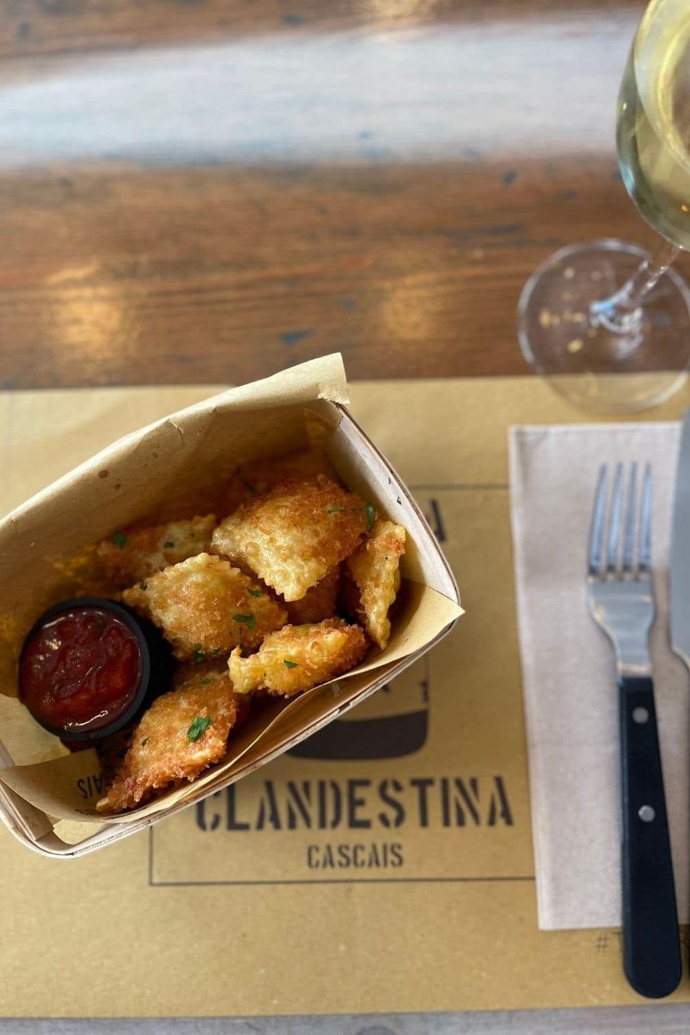 Close-up of crispy fried ravioli in a takeout box served with tomato sauce, next to a half-empty glass of white wine, on a wooden table with a branded napkin saying 'Clandestina Cascais