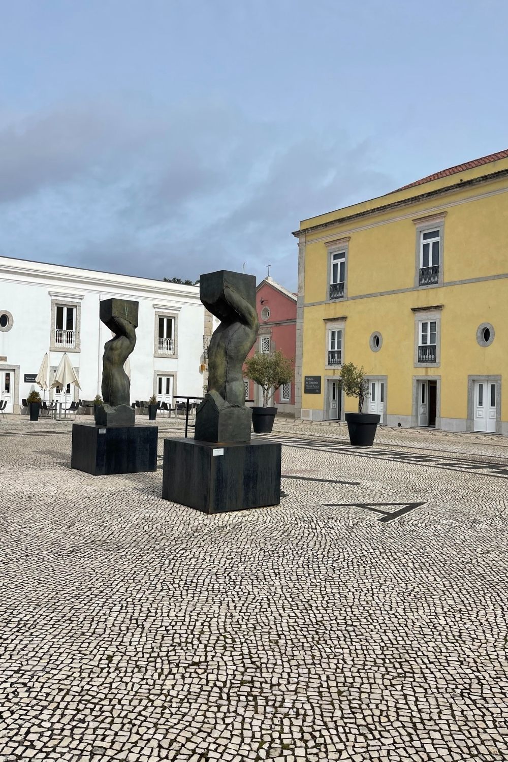 Three abstract sculptures stand in a public space, each with a distinct twisted form, possibly representing figures or movements. The sculptures are set against the background of classic European architecture and a cobblestone square, suggesting an area rich in history and culture.