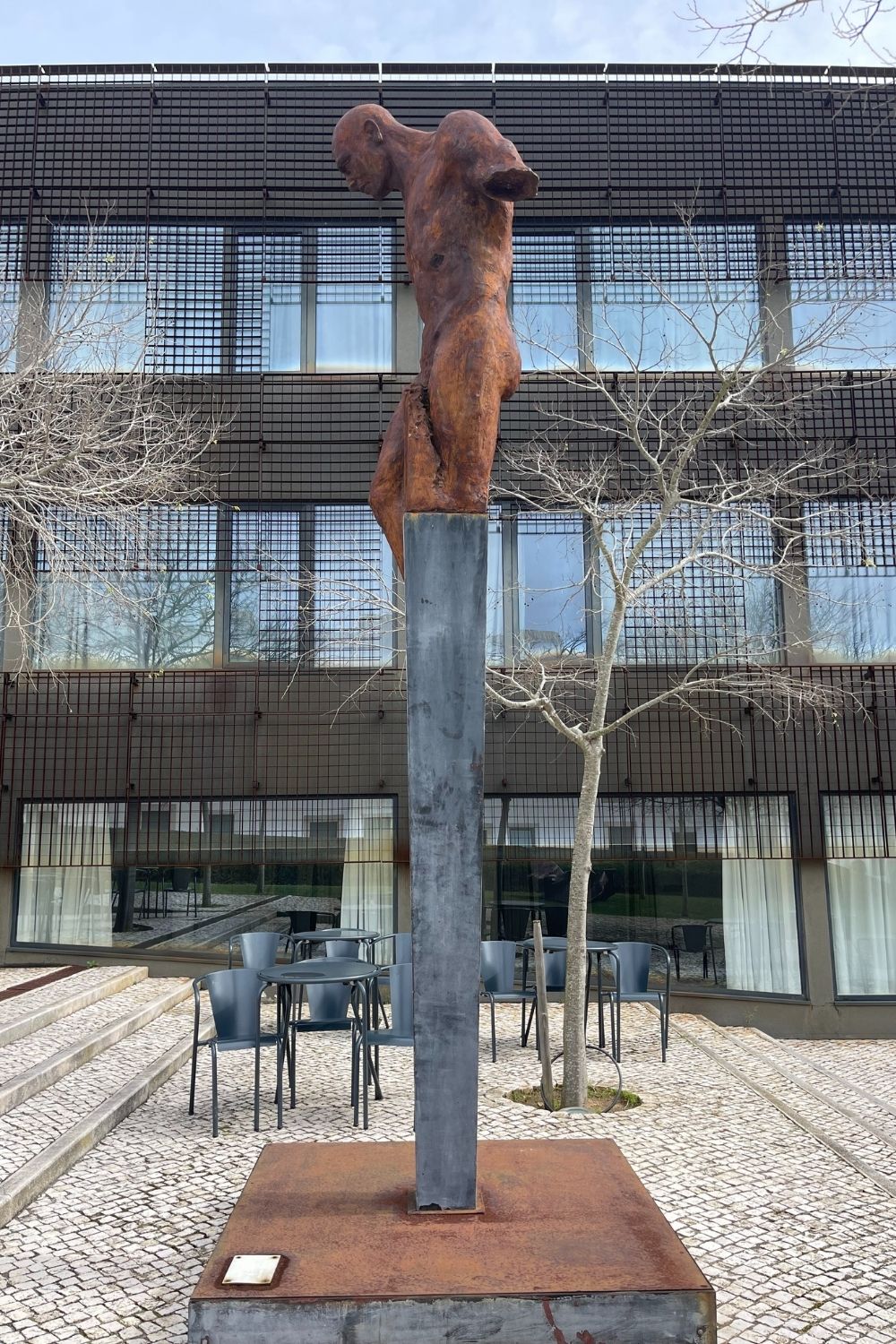 A rust-colored sculpture of a figure atop a tall steel pillar is placed before a modern building with a grid facade. The sculpture appears contemplative or in mourning, adding a touch of emotion to the otherwise urban and architectural environment.