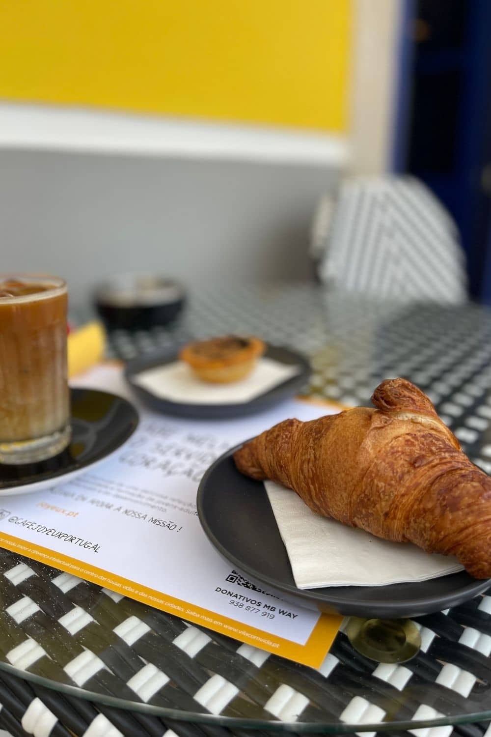 A table with a blurred background emphasizes the items in the foreground: a glass of iced coffee, a pastel de nata on a plate, and a fresh croissant. The focus on the delicious snacks and the use of a narrow depth of field suggest a casual, enjoyable moment, likely part of a leisurely café experience.