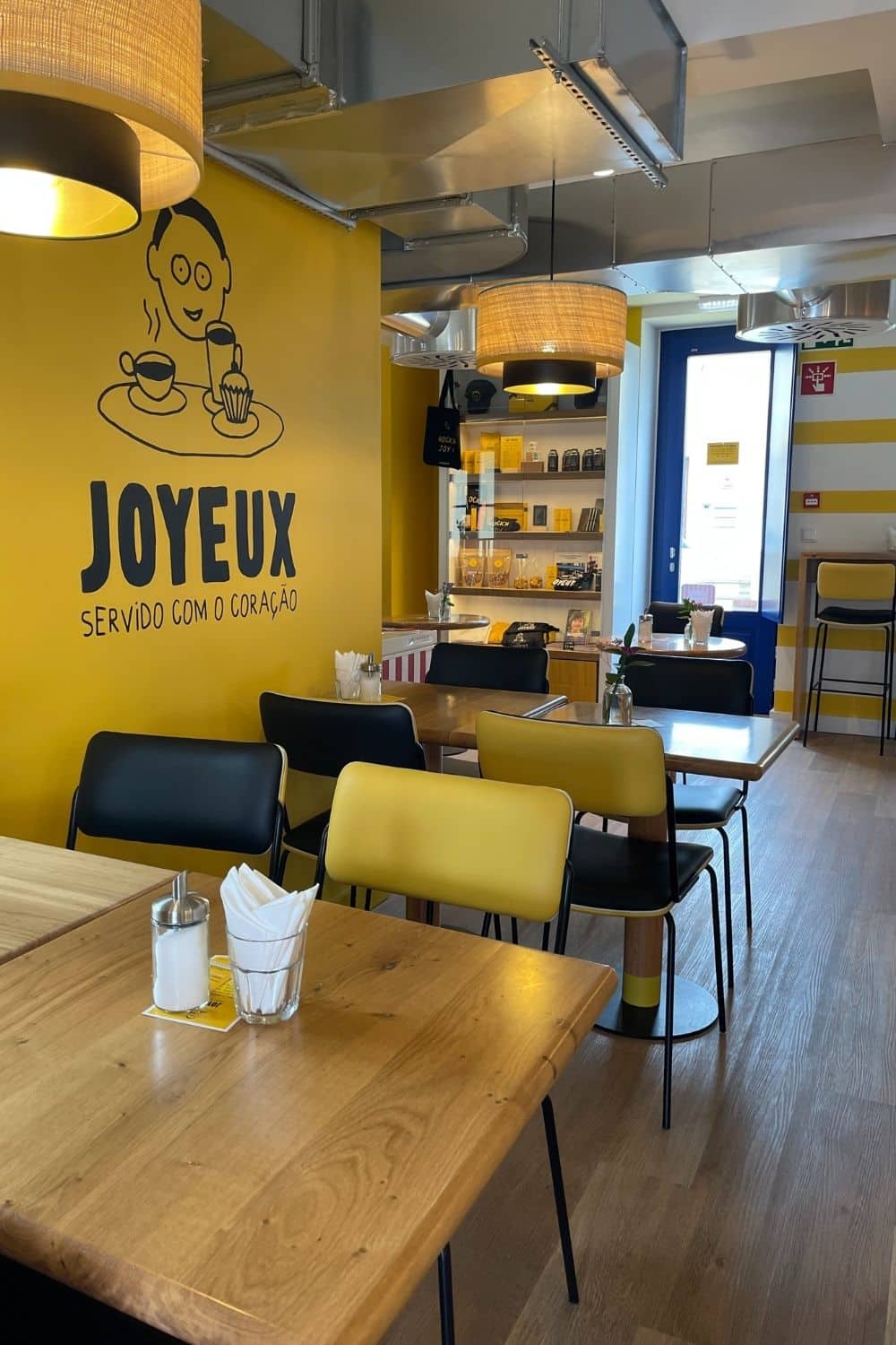 Inside a café with a warm and inviting ambiance. The interior is brightly lit with natural light. The yellow walls are adorned with a simple, playful drawing of a man enjoying coffee and a dessert, accompanied by the café's slogan "SERVIDO COM CORAÇÃO" (Served with heart). Wooden tables with yellow and black chairs are neatly arranged, and the counter area displays an assortment of goods.