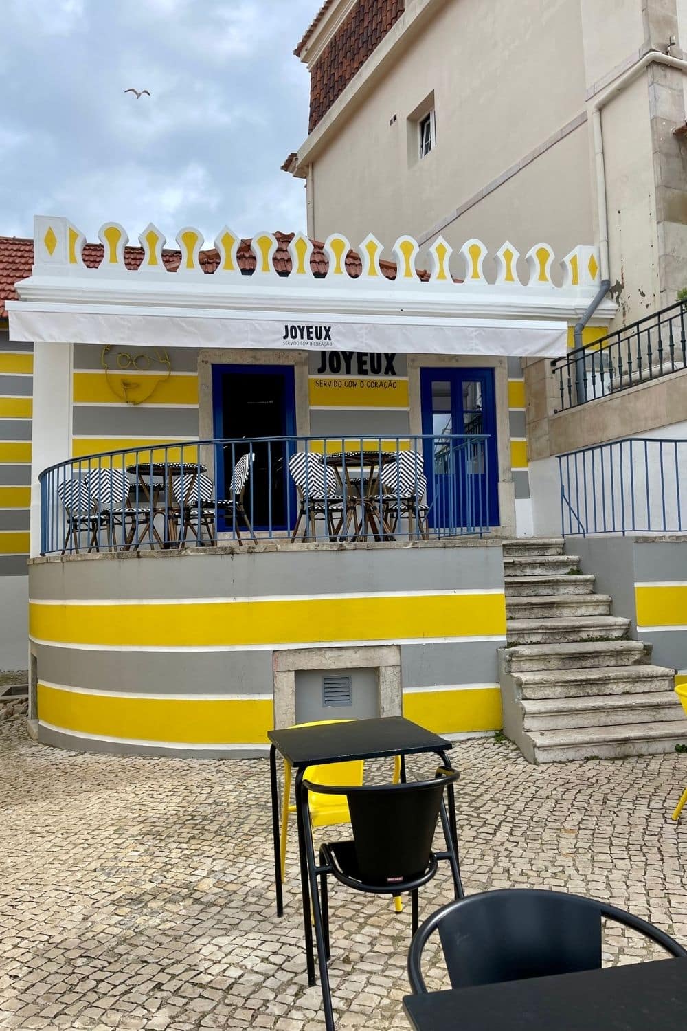 A charming café exterior is painted in bright yellow and white stripes, with a bold blue sign reading "JOYEUX" above the entrance, implying a cheerful, welcoming atmosphere. The terrace has several tables with patterned tablecloths, and the ground is traditional cobblestone. A simplistic line drawing of a smiling face adorns the wall beside the café's name, enhancing the joyful vibe of the establishment.