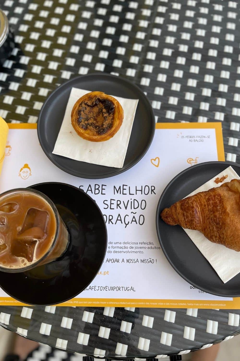 A close-up of a cafe table with a black and white geometric pattern. On it is a traditional Portuguese pastel de nata on a small plate, a croissant, and a glass of iced coffee. Below the coffee is a paper placemat with the text "SABE MELHOR SERVIDO COM CORAÇÃO" (Tastes better served with heart), reflecting the cafe's ethos, and there is a card indicating that orders are placed at the counter.