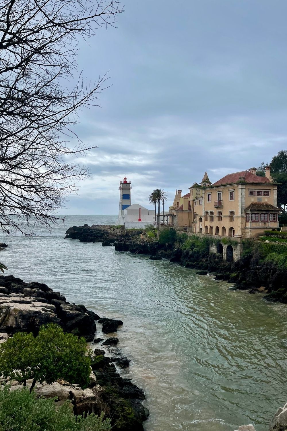 A picturesque view of Cascais's rocky coastline with a lighthouse and classic European architecture in the background, under a dramatic overcast sky, portraying a blend of natural beauty and historical charm.