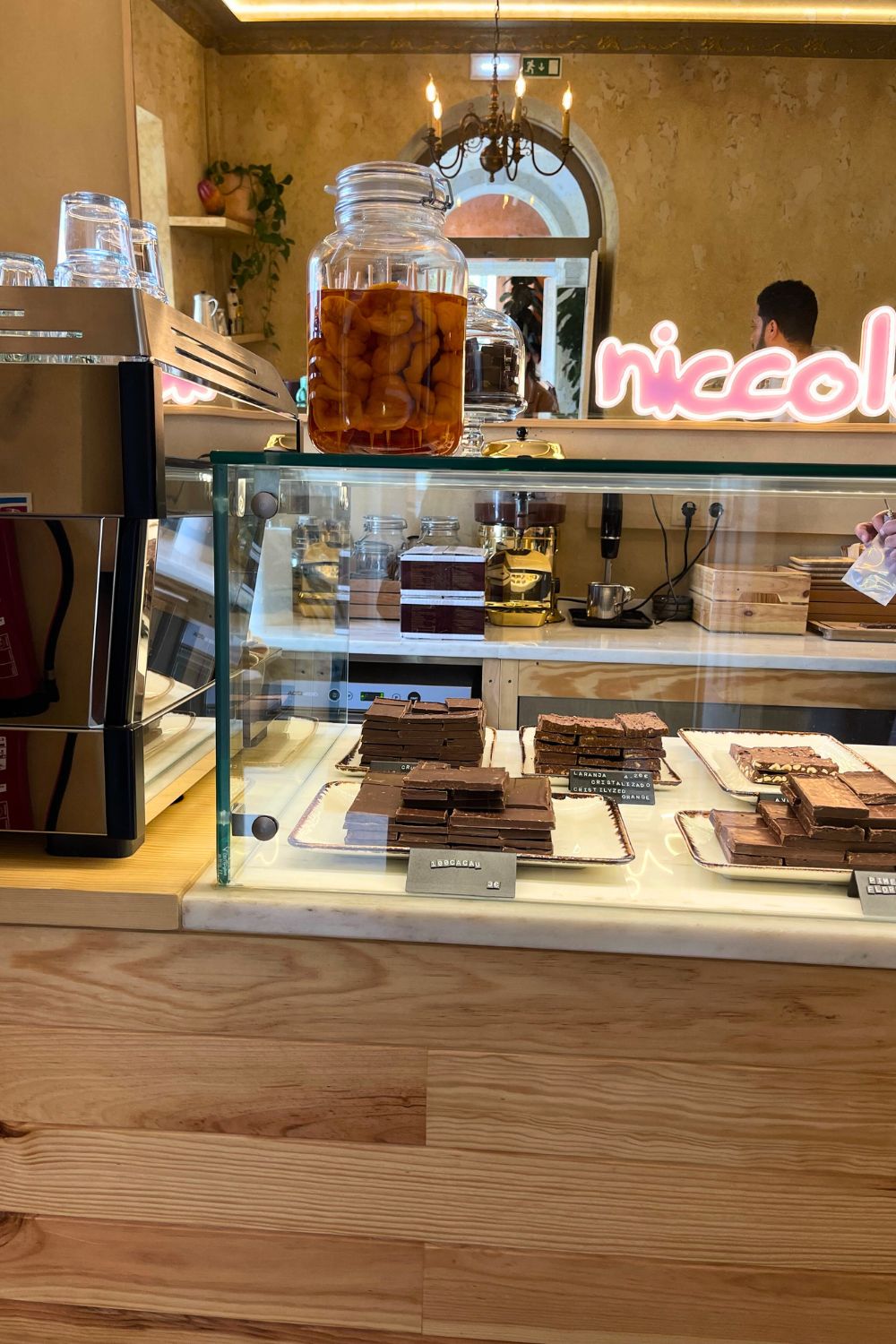 The interior of Niccolò café showcases a selection of fine chocolate bars neatly presented in a glass display case, with warm lighting and a stylish neon sign reflecting in the mirror, creating a cozy and upscale atmosphere for chocolate enthusiasts.