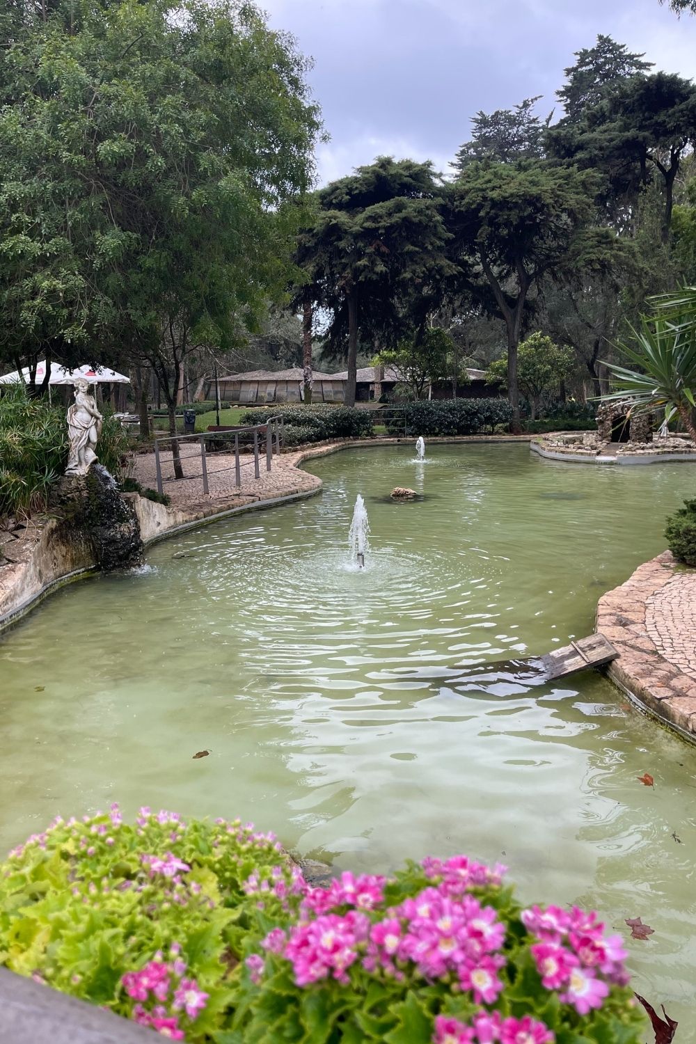 This image captures a classical garden scene featuring a green pond with a fountain, surrounded by trees, flowers, and a classical statue, contributing to an air of elegance and tranquility. The overcast sky suggests a peaceful, contemplative environment, ideal for a leisurely stroll.
