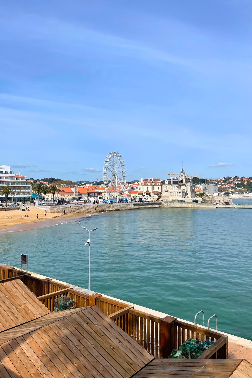 A serene waterfront view in Cascais, Portugal, with a picturesque Ferris wheel towering over the beach and town. The clear blue sky is reflected in the tranquil waters, and the wooden deck in the foreground adds a warm, inviting touch to the scenic coastal panorama.