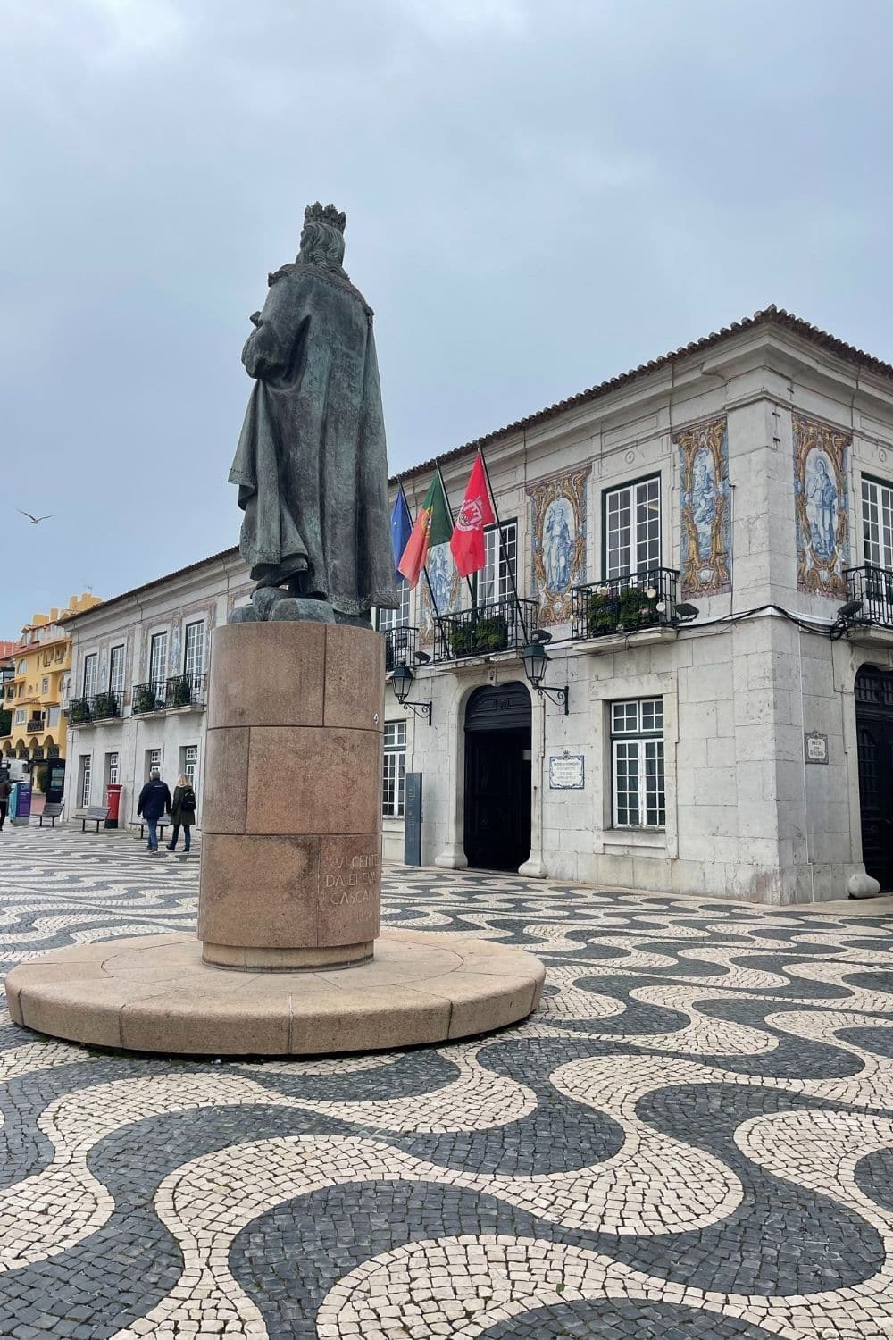 A historical bronze statue of a robed figure, presumably a significant local figure, on a stone pedestal, with an intricate mosaic pavement below. The statue is in a public square flanked by buildings with traditional Portuguese azulejo tiles and flags