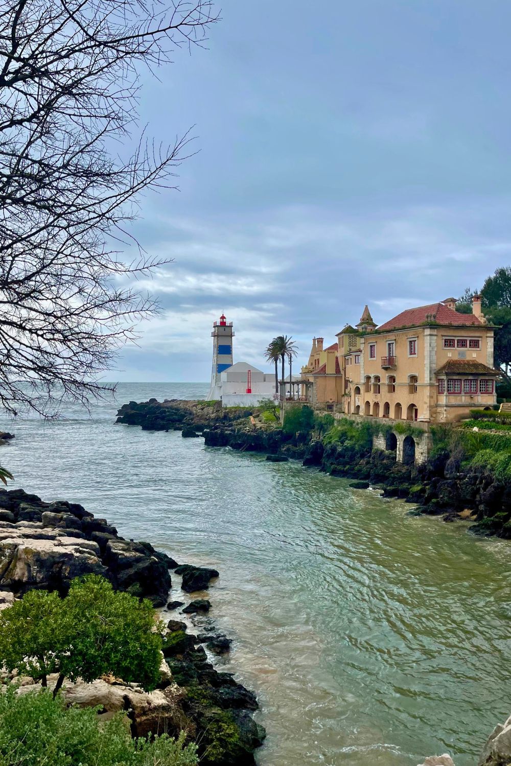 A serene scene at the Santa Marta Lighthouse in Cascais, Portugal, with a view of the beacon overlooking a calm sea beside a historic building, framed by bare branches against an overcast sky.