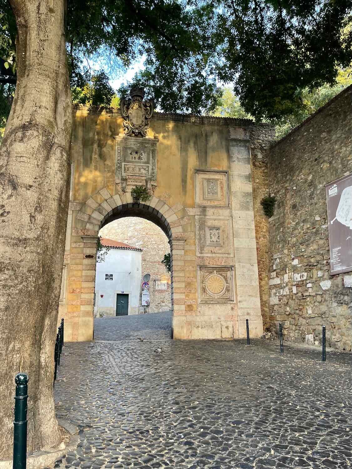 Ancient arched gateway adorned with stone carvings, leading into the shaded, cobbled streets of Lisbon's historic district