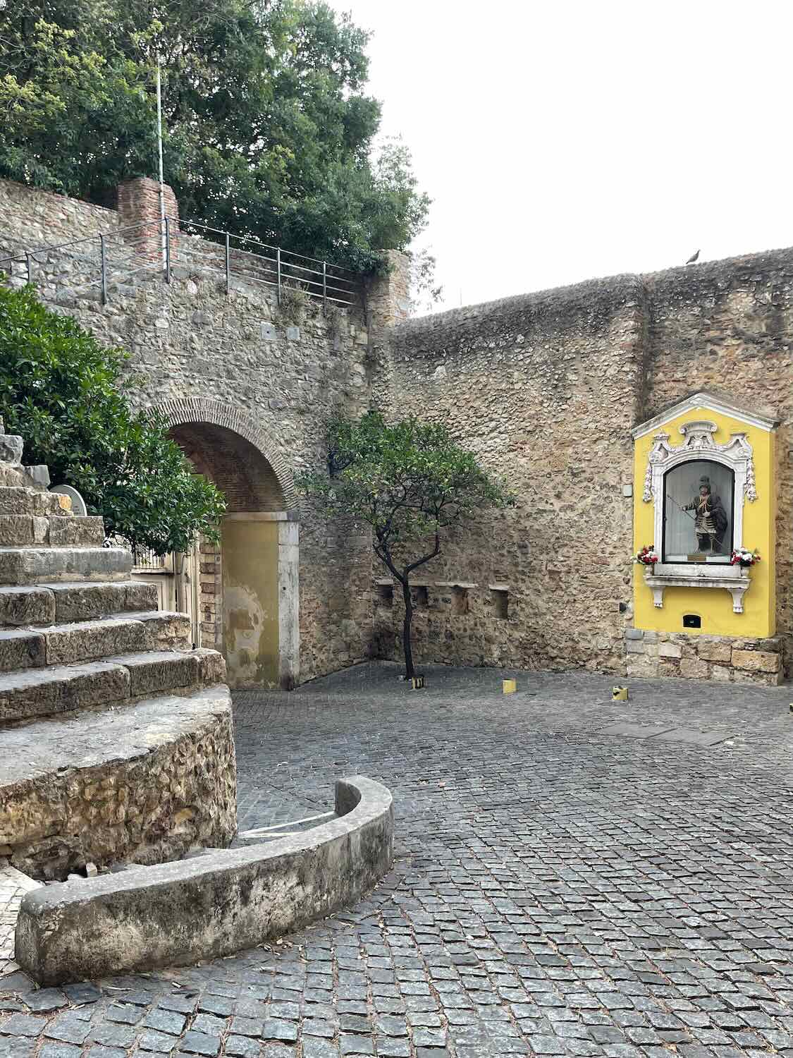 A quiet courtyard with a cobblestone pathway leading to a small, ornate shrine built into the yellow walls of Lisbon's Castle.