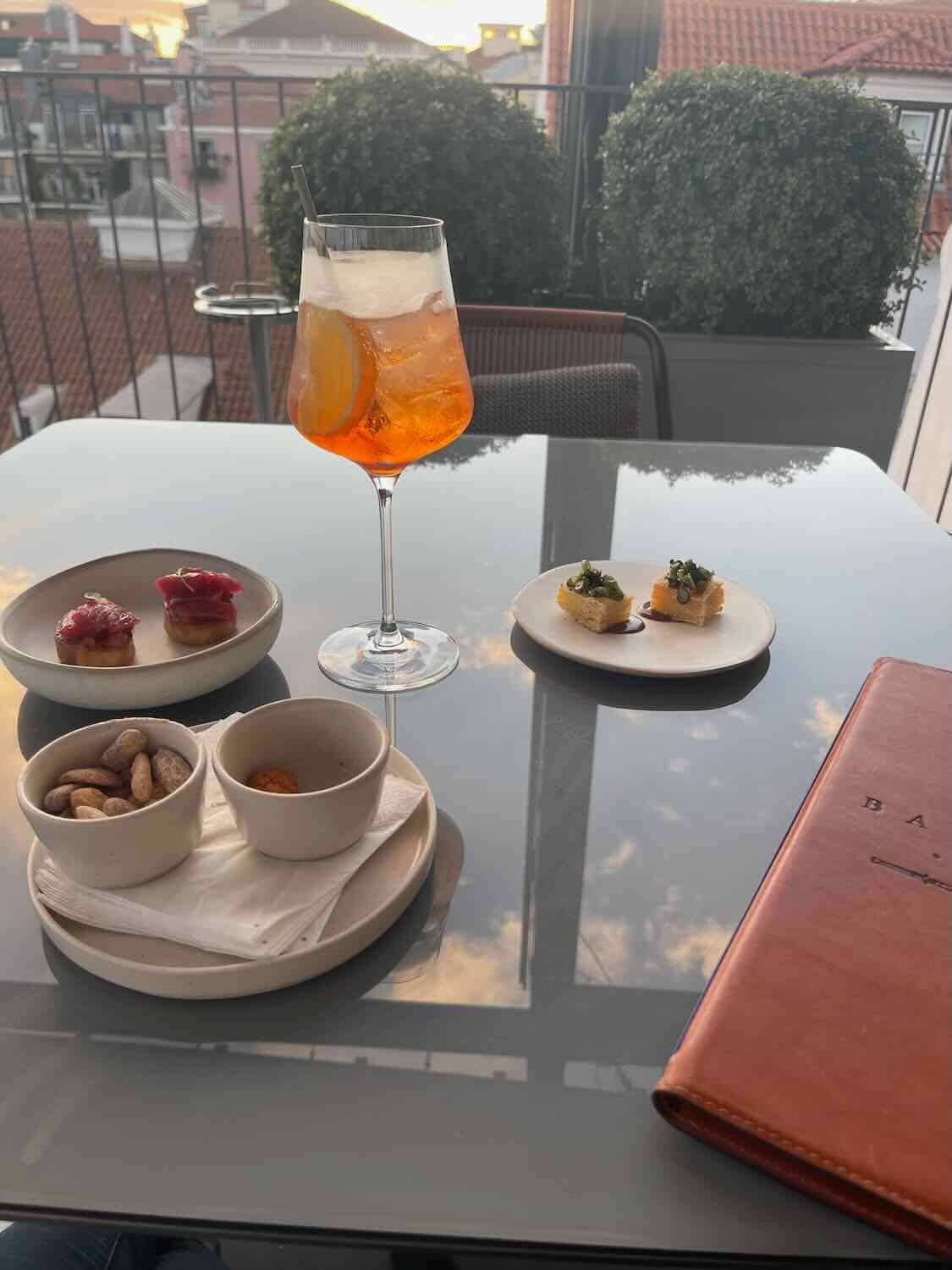 A refreshing Aperol spritz and a selection of gourmet appetizers on an outdoor terrace with a view of Lisbon's charming rooftops at dusk.
