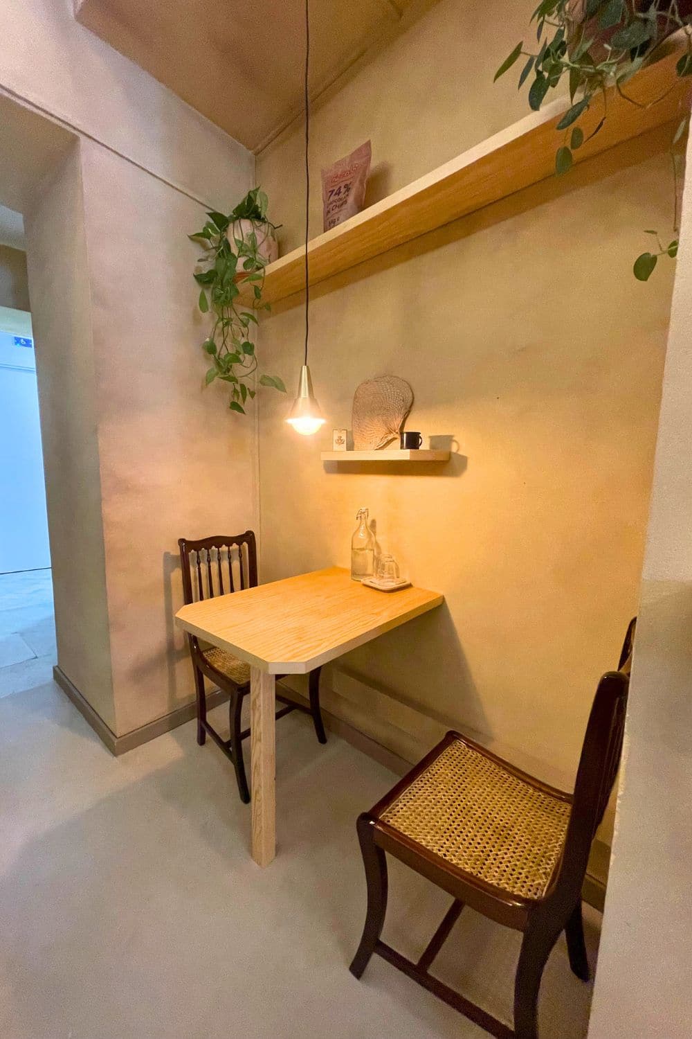 A quaint corner inside Niccolò café, radiating a serene ambiance with a single wooden table and a woven chair. A hanging plant and a warm pendant light above create a cozy, intimate space for savoring coffee and chocolates in peace.