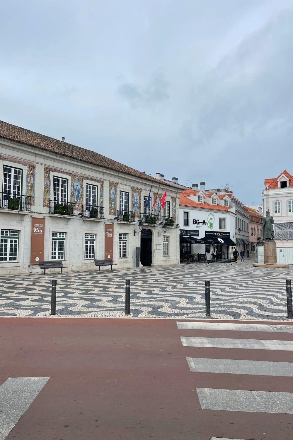 View of a historic square in Cascais with traditional Portuguese pavement design and a row of buildings adorned with azulejo tiles, under a cloudy sky, evoking the charming European coastal town atmosphere.