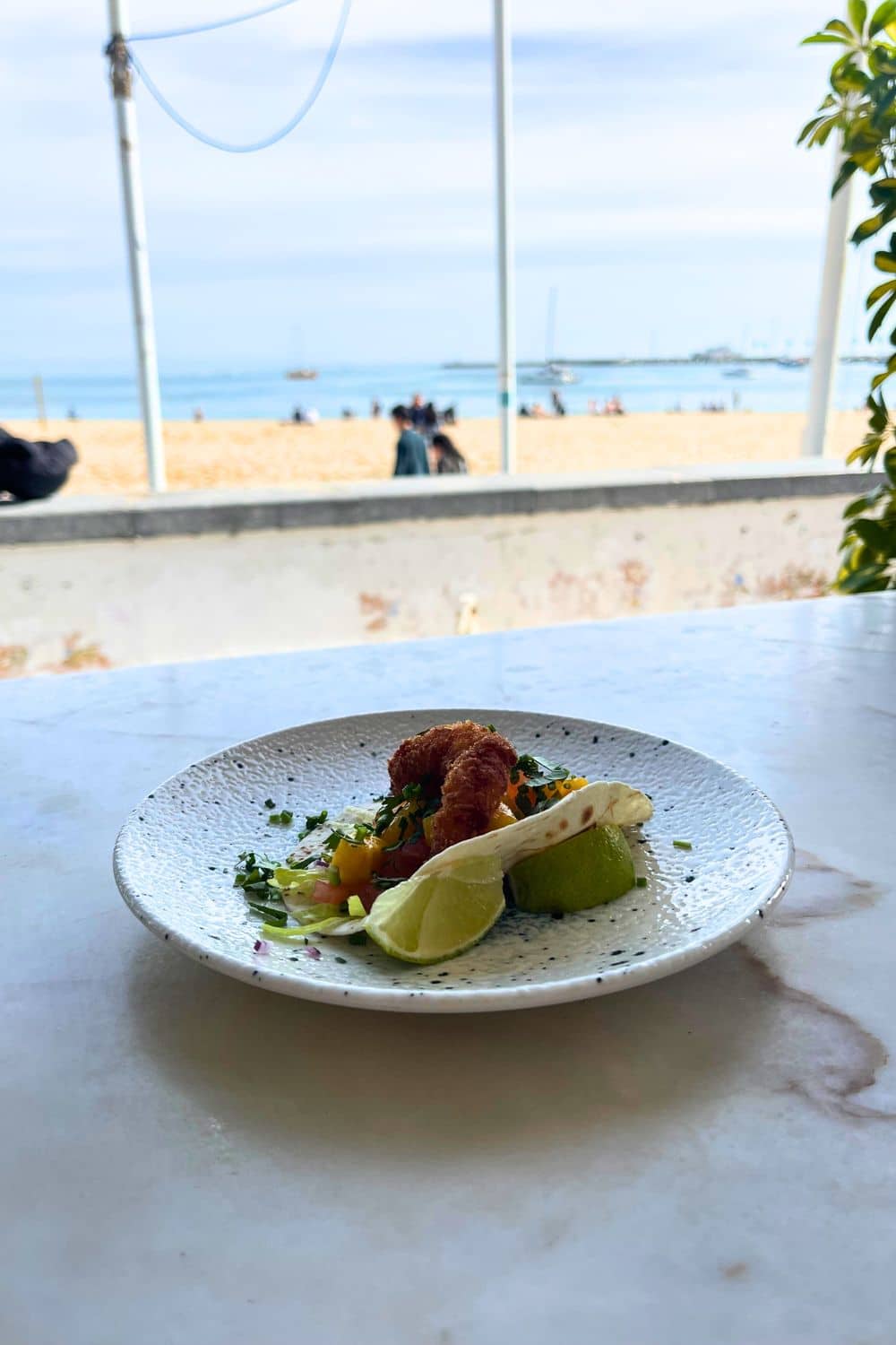 A plate with a breaded shrimp taco garnished with fresh lime on a speckled ceramic plate, set on a marble table with a blurred beach scene in the background, offering a dining experience with a view in Cascais.