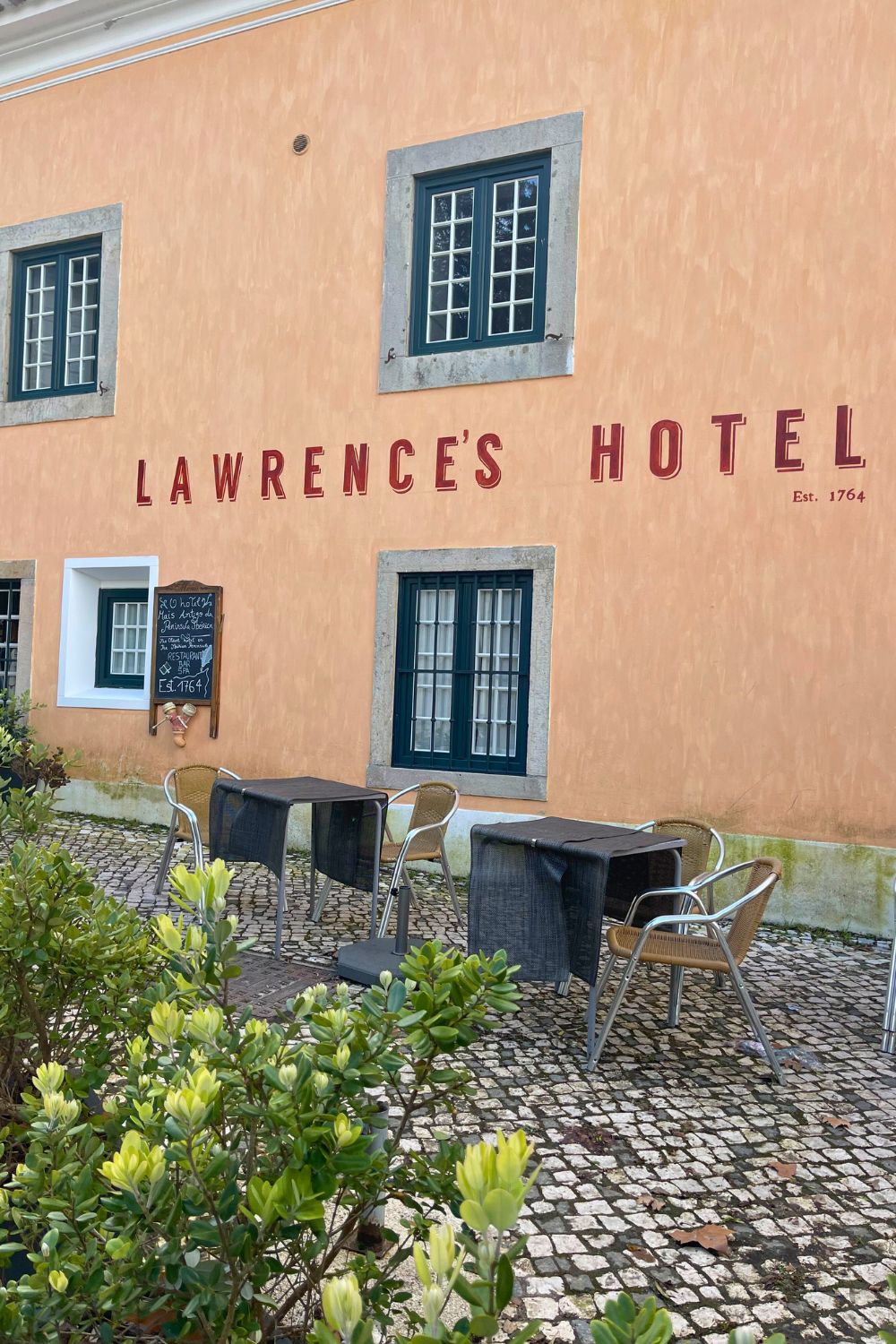 The peach-colored facade of Lawrence's Hotel with the name and 'Est. 1764' in red lettering, complemented by traditional grey-framed windows, and a blackboard displaying the hotel's history. A cobblestone courtyard with outdoor seating invites guests to relax outside this historical establishment.