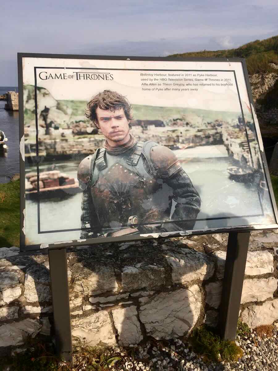 Informational display board for 'Game of Thrones' at Ballintoy Harbour, known as Pyke Harbour in the show, featuring Alfie Allen as Theon Greyjoy in armor. The background shows a picturesque view of the harbor, blending the fictional world with the real-life filming location.