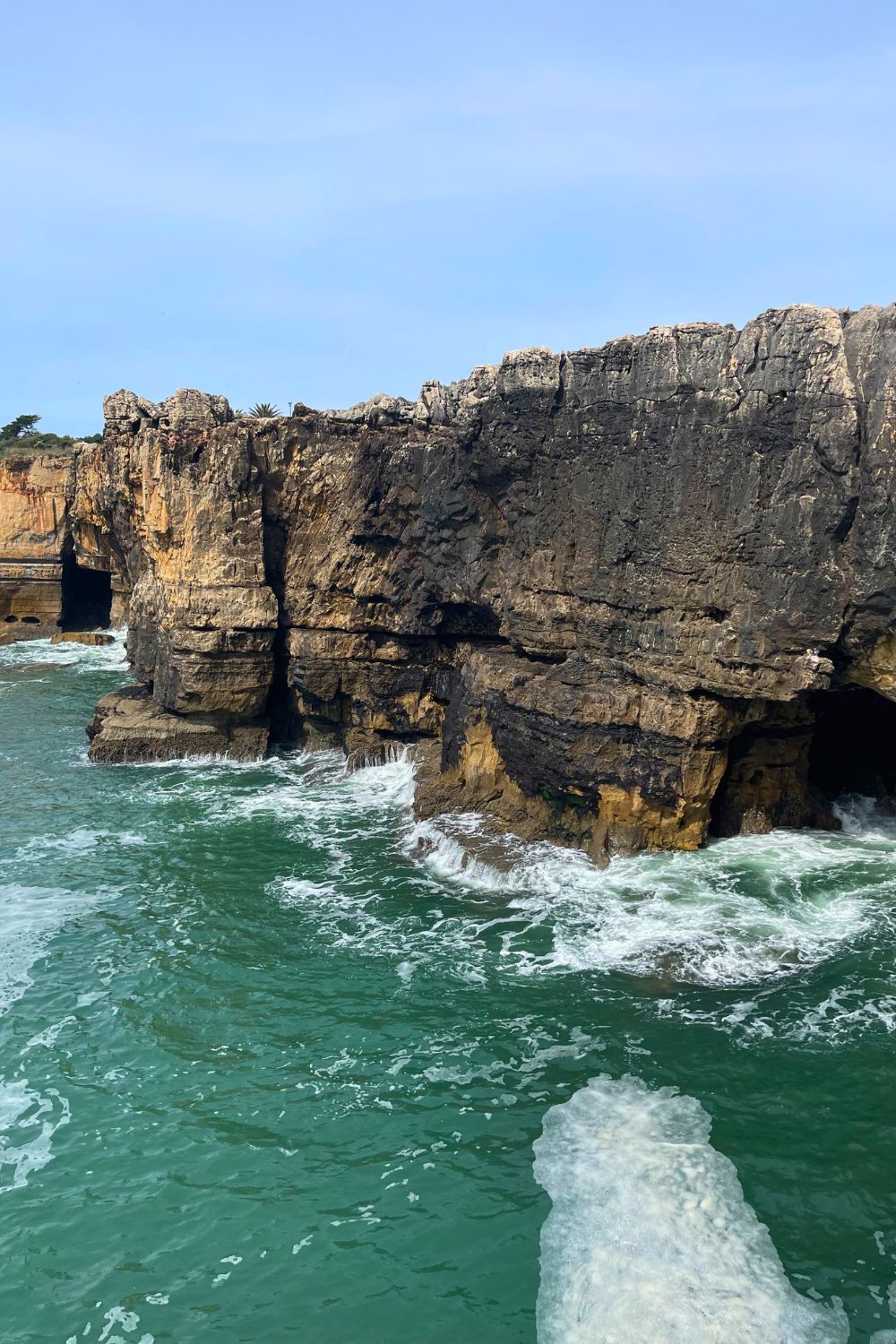 A closer perspective of Hell's Mouth (Boca do Inferno) in Cascais, showing the dramatic rock structures and the powerful waves crashing into the caverns. The clear emerald waters and the stratified rock faces create a majestic natural spectacle, highlighting the raw beauty of Portugal's coastline.