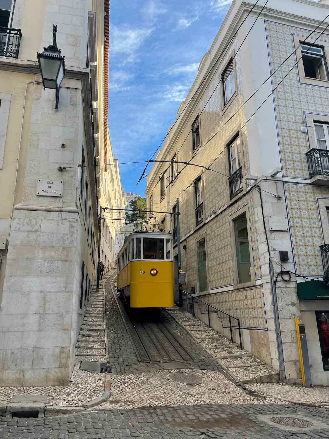 A classic yellow tram ascends a steep, cobbled street in Lisbon, framed by traditional tiled buildings under a clear blue sky, capturing the essence of the city's historic charm.