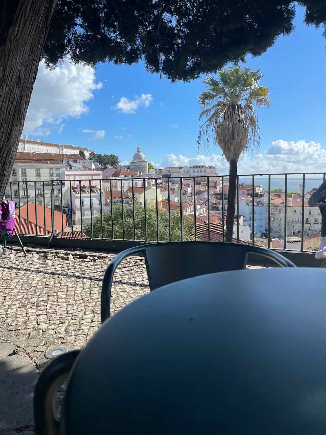 View from a café terrace in Lisbon with a foreground of a table and chair, offering a serene vista over the city's red rooftops, with the National Pantheon's dome prominent in the background, under a sunny sky with fluffy clouds.