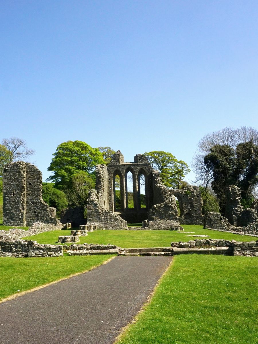 Sunny day at Inch Abbey, with its ancient stone ruins standing among vibrant green grass, a clear blue sky above, and a paved pathway leading towards the historic site.