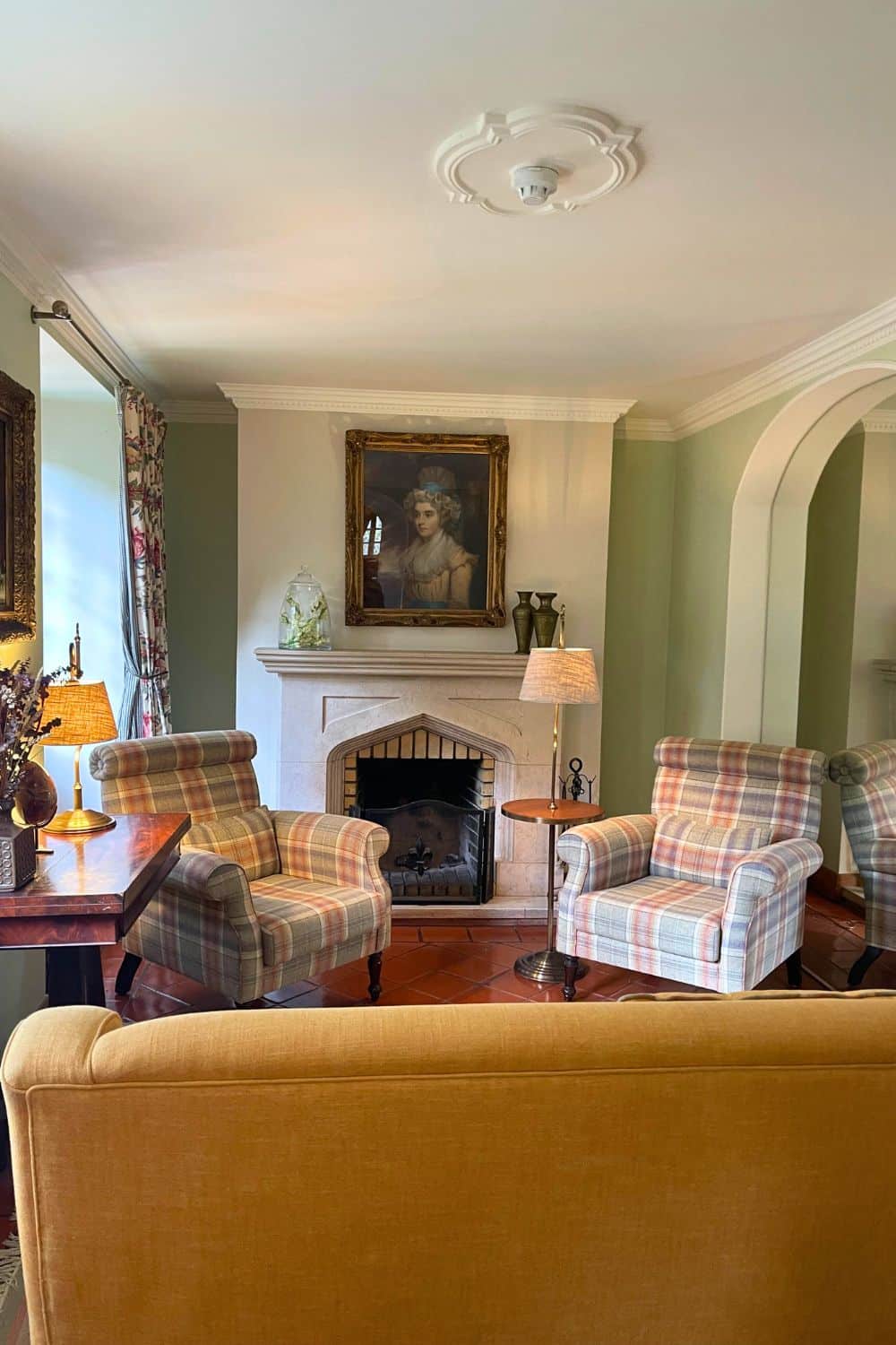A cozy interior space within Lawrence's Hotel featuring a classic fireplace with an ornate painting above it, flanked by plush, patterned armchairs and an inviting golden sofa, all under warm lighting that highlights the room’s elegant decor and welcoming ambiance.