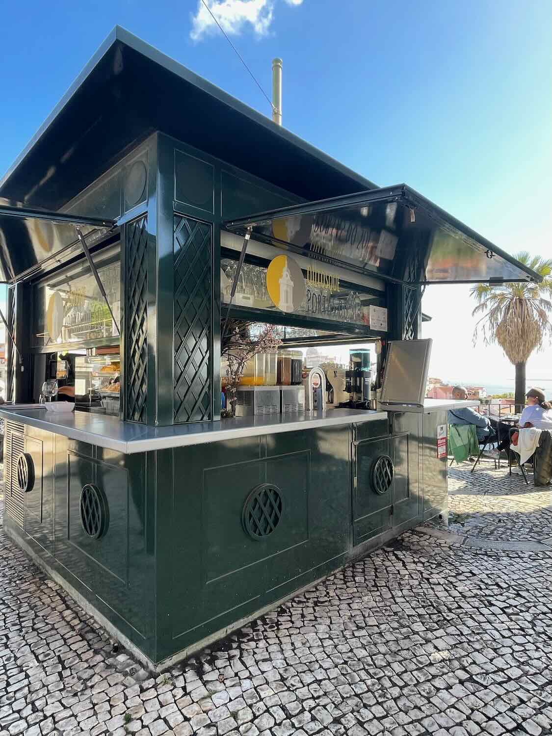 A quaint, green-painted kiosk cafe in Lisbon under a clear sky, offering a spot for refreshing drinks and a casual outdoor seating area