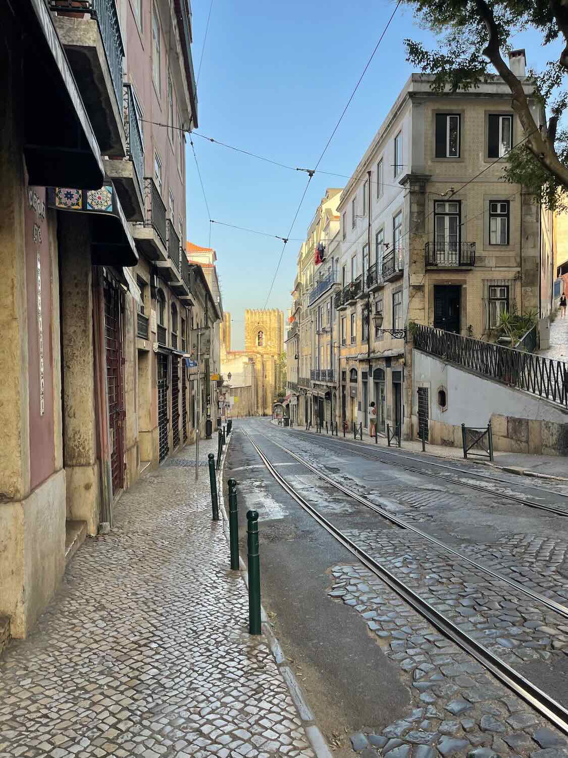 Deserted cobblestone streets in the early morning light with Lisbon's traditional architecture and tram lines leading towards the Se Cathedral in the distance.