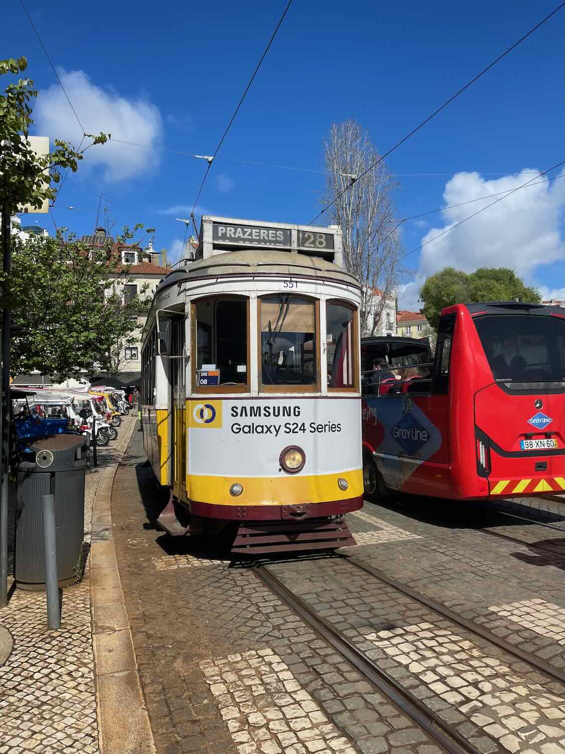 Lisbon's iconic Tram 28, adorned with a Samsung advertisement, waits for passengers against a backdrop of bright blue skies and city foliage.