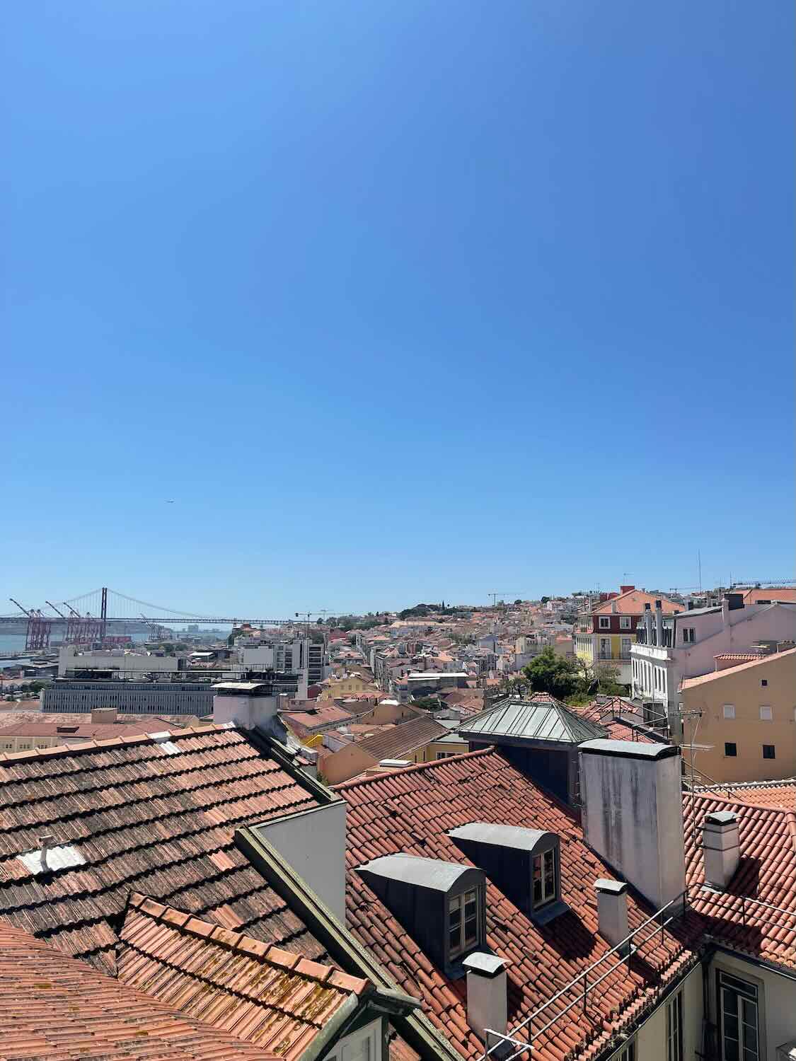 A rooftop view over Lisbon's terracotta roofs, with the 25 de Abril Bridge in the distance, under a clear blue sky, showcasing the city's topography.